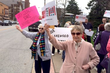 A group of concerned Lakewood, Ohio, residents, including Ann Allen, front right, and Marie Birsic, left, gather in April outside the Lakewood Hospital near Cleveland to protest its closure. “Lived here for 44 and half years, we’ve all used it and we’ve got to do this,” Birsic says. (Photo by Sarah Jane Tribble/WCPN)