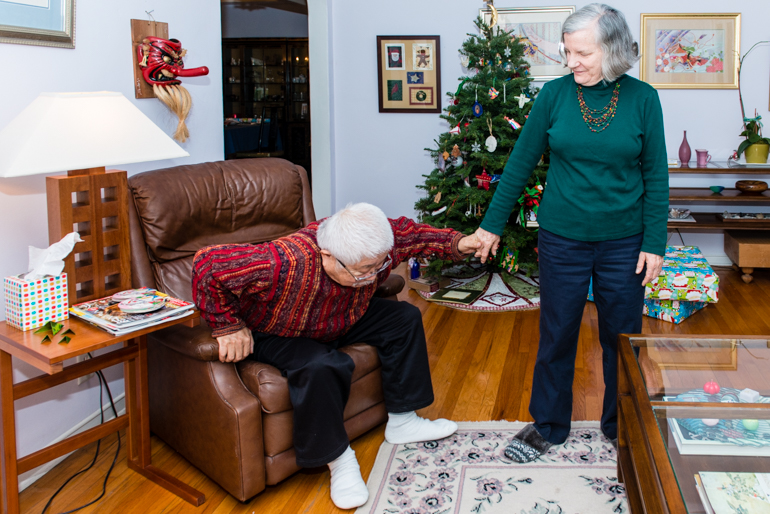 Diana Matsushima helps her husband Yoshi up from the recliner chair. After her husband suffered a stroke, Matsushima needs to help him get around. (Heidi de Marco/KHN).
