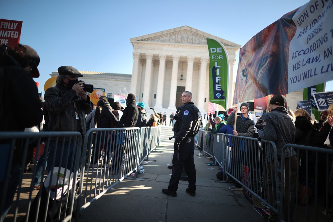 Two metal fences separate two sides of protesters standing in front of the U.S. Supreme Court. Anti-abortion protesters on the right hold signs and wave banners with sayings like "DE for life." Pro-abortion advocates stand on the left. One holds a sign that reads, "Liberate abortion." A police officer stands between the two fences.