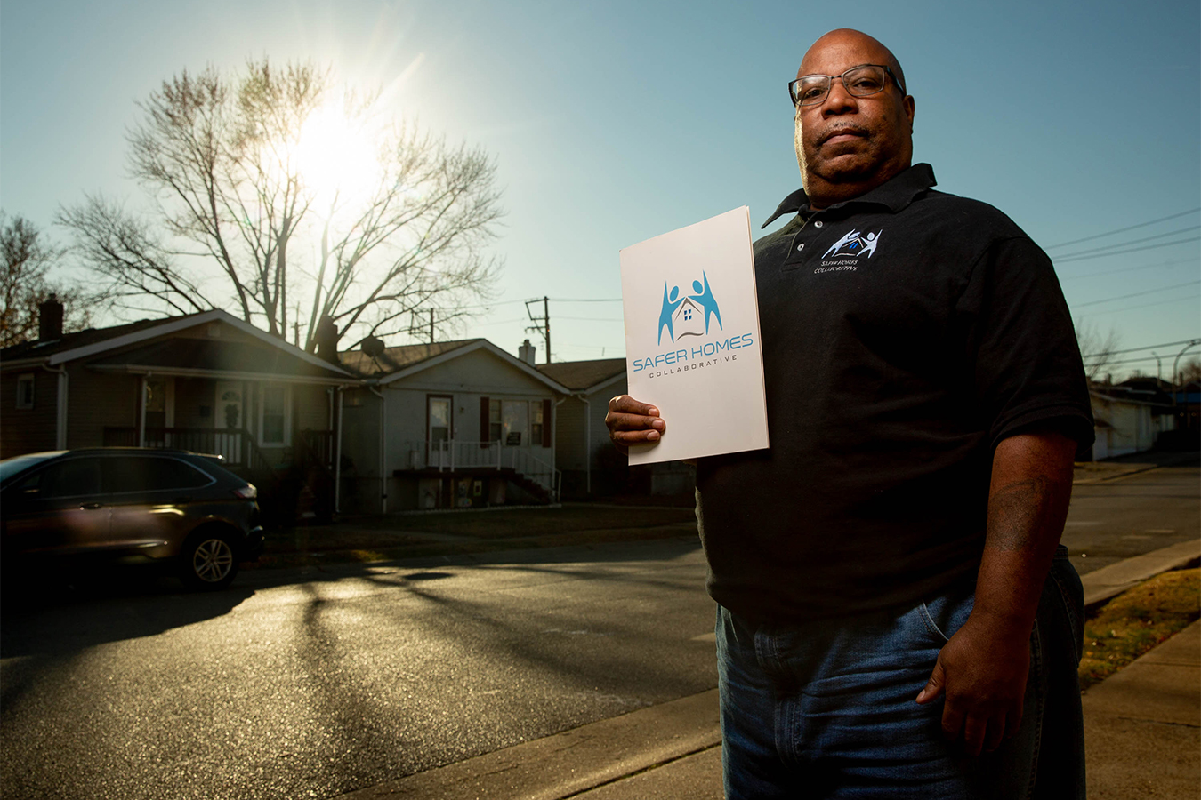 Bill Mays stands for a portrait on the sidewalk, holding a folder with the logo and name of his employer, Safer Homes Collaborative. Warm sunlight hits from the left side, leaving long shadows.