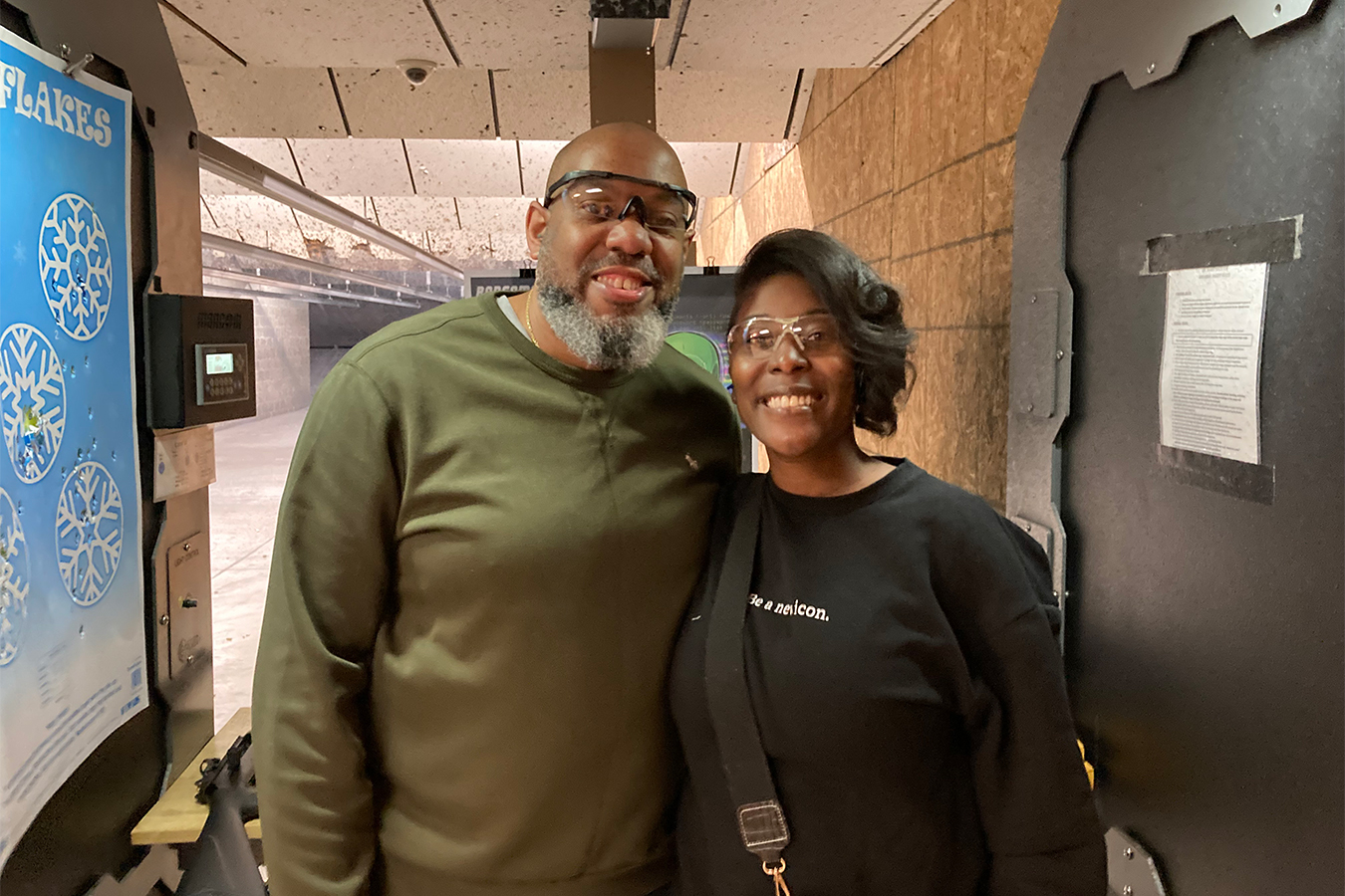 Sharis and Russell Lewis pose for a photo together at the gun range, standing next to each other, smiling. They are both seen wearing protective glasses.
