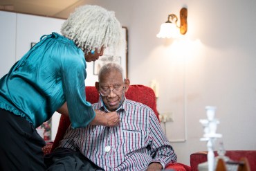 On the left side of the image, a Black woman with short, white dreadlocks leans over a senior man, her father, to help button his shirt. The woman wears a turquoise shirt, while the man sits in a red recliner. They are in their home living room.