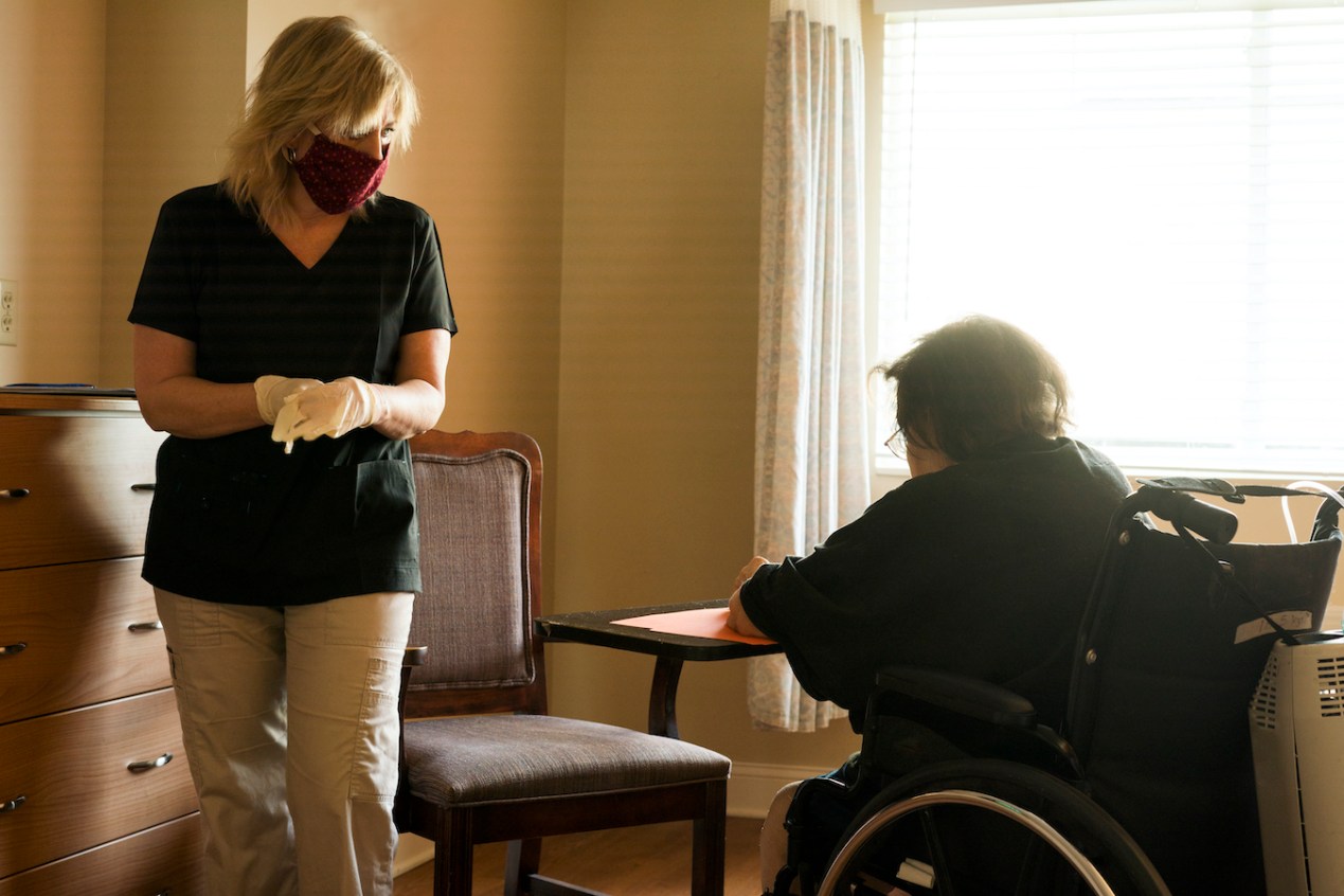 Blond female healthcare worker wearing scrubs, gloves and a red fabric face mask approaches a patient on oxygen sitting in a wheelchair to begin a speech therapy session amid the corona virus COVID-19 pandemic.