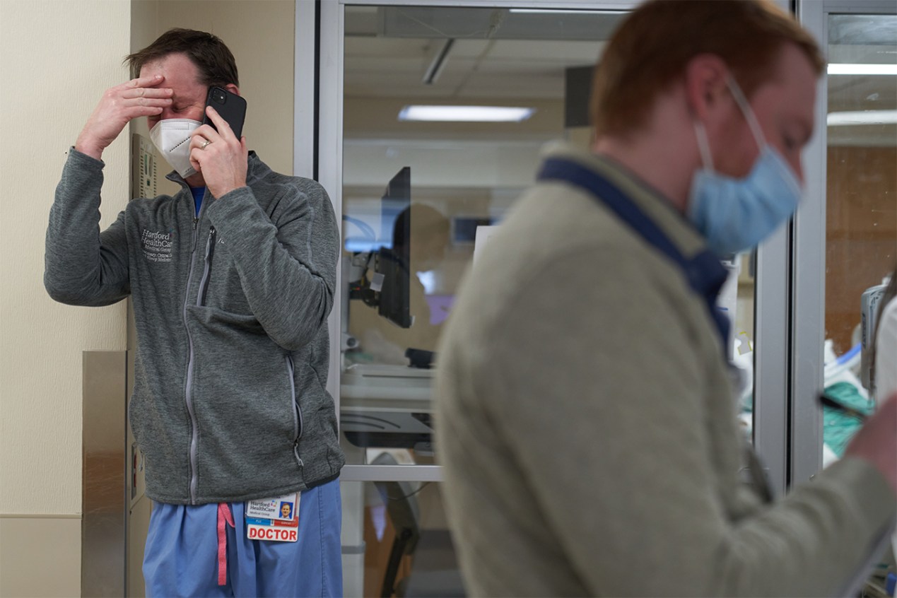 A doctor wearing a jacket over his scrubs presses his hand to his head and winces while talking on the phone in a hospital. Another man is seen out of focus on the right in the foreground.