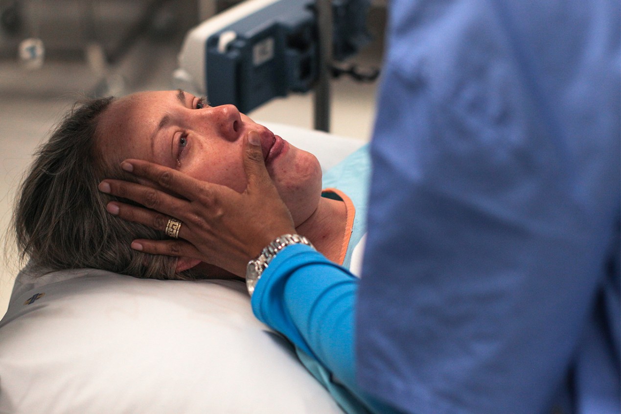 A woman is seen lying down in an operating room, a tear in her eye. A doula holds a hand to her cheek.