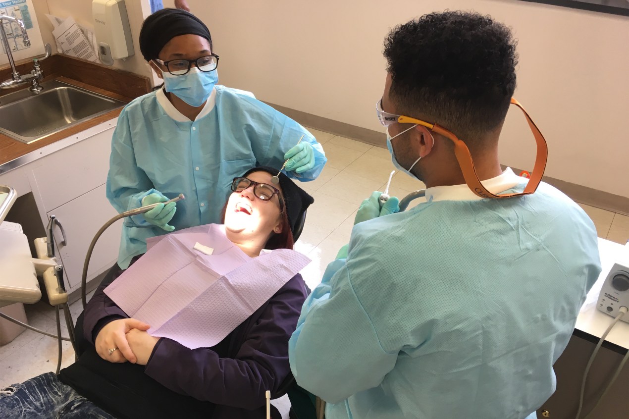 Mary Ashlee Tosh lies in a dental chair while Dr. Ratrice Jackson sits to her side, holding dental tools in both of hands. A man is seen in the foreground in the left of the frame.
