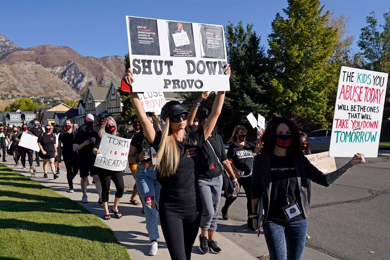 Paris Hilton leads a line of protesters dressed in all black. She holds a sign that reads, "Shut down Provo." Others hold signs that read, "Torture is not treatment," and "The kids you abuse today will be the ones that will take you down tomorrow."