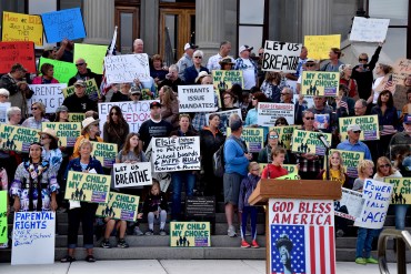 A crowd holding signs stands on the steps of the Montana capitol. The signs display anti-mask slogans: "Let us breathe," and "My child, my choice."