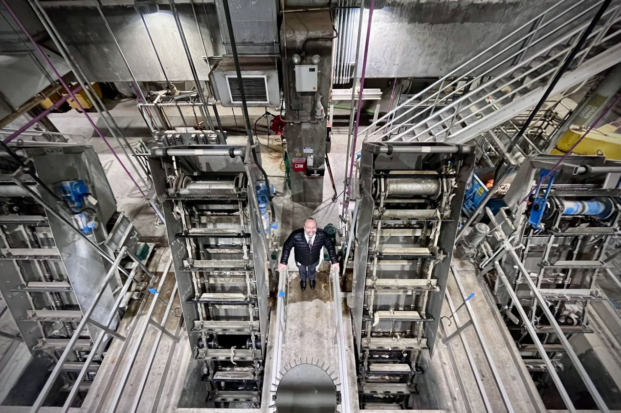 Pieter Van Ry is seen standing between two machines, smiling at the camera. He is seen from above, and a wide angle lens shows the facility around him.