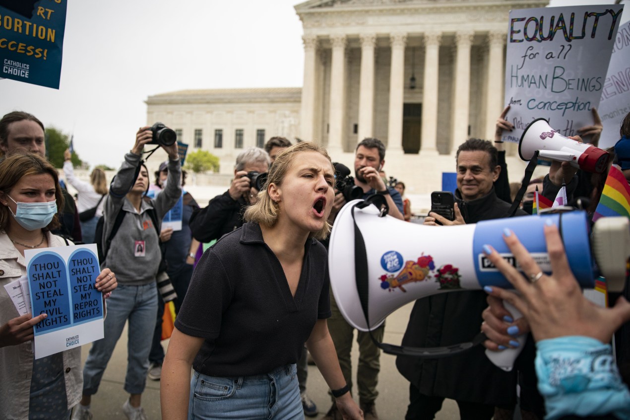 A photo shows a woman protester in front of the Supreme Court yelling at someone off camera. A crowd of photographers is taking photos behind her.