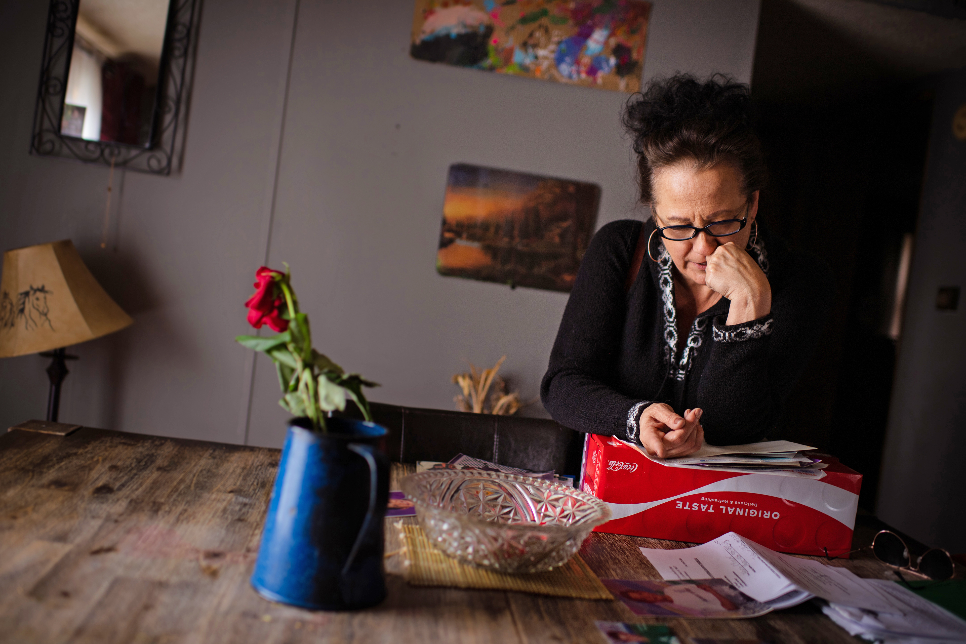 Marla Ollinger is seen looking at papers and photos on a table in her home. Light is coming in from the left, casting the right side of her in shadow.