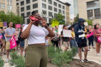 Tia Freeman is seen cheering at a protest, cupping her hands around her mouth. The background shows blurred figures holding signs.