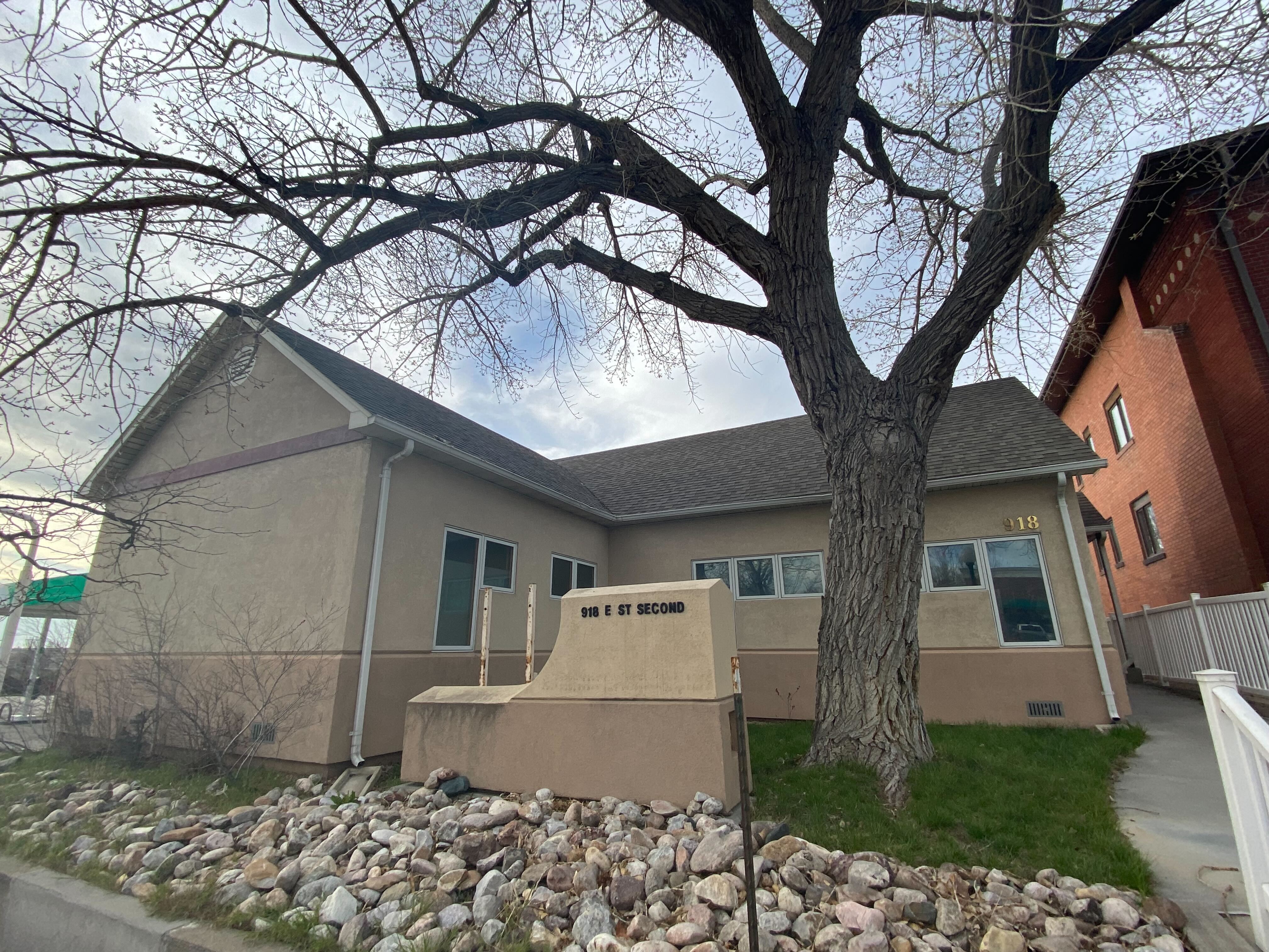 A photograph of the Wellspring Wyoming Health Access clinic. The single-story building is a khaki brown color, and a has a large tree growing in the small yard in front of it.
