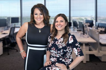 Ruby Gadelrab (left) stands next to Katie Coleman (right). They both are smiling widely at the camera. The background shows their office, out of focus.