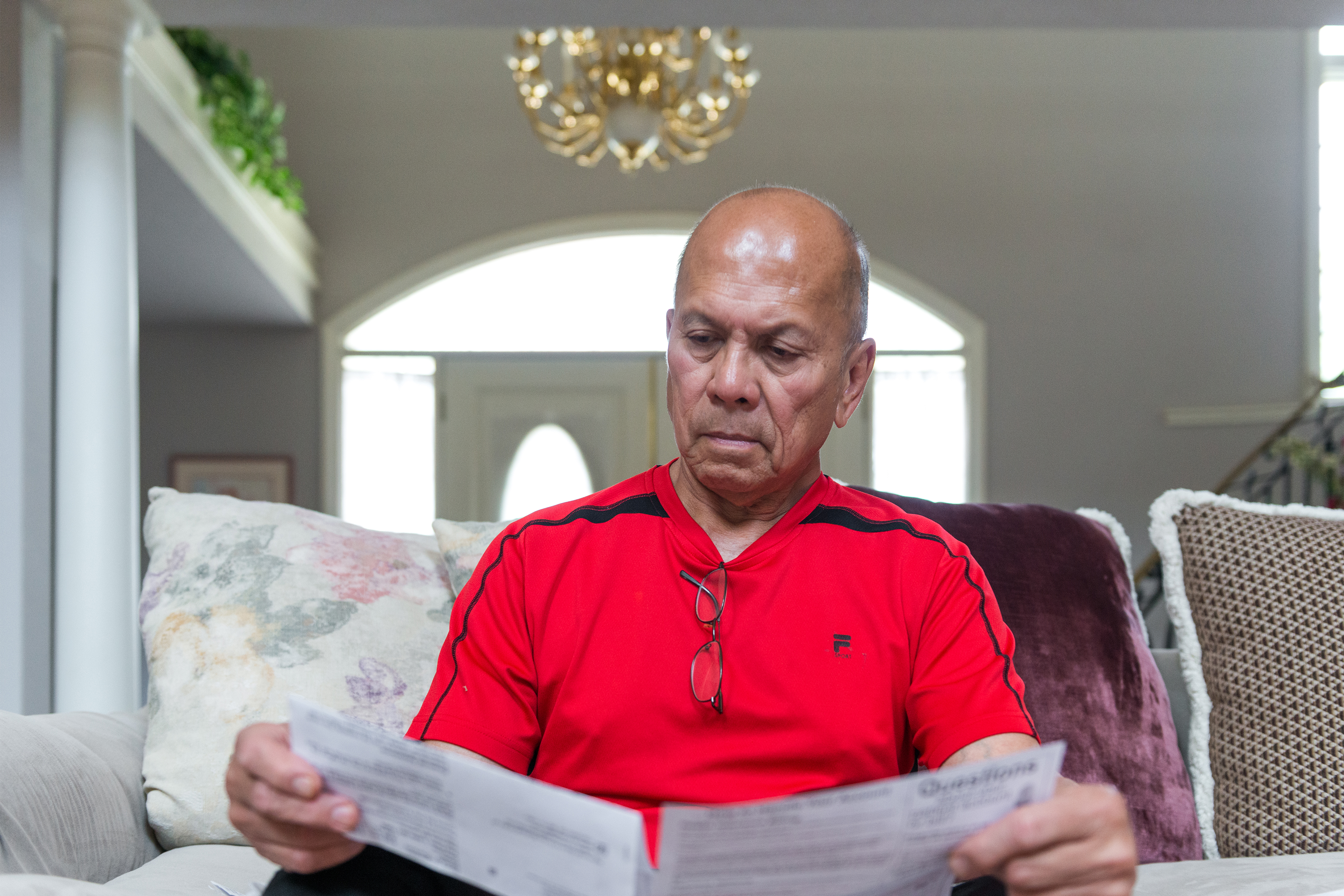 A photo shows Danilo Manimtim sitting on a couch at home looking at medical bills.