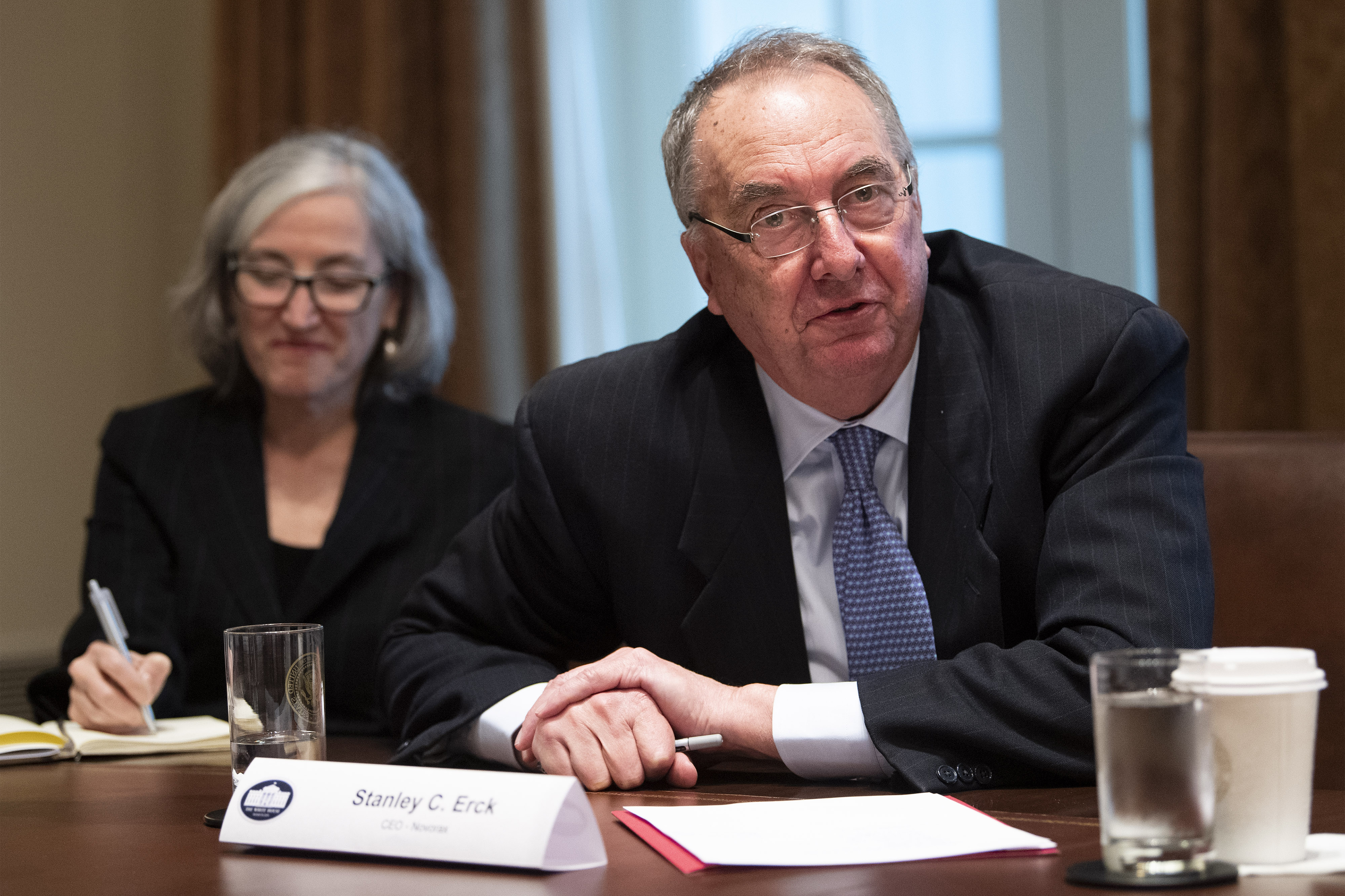 Stanley Erck is seen speaking at a table, with a woman blurred in the background behind him. A nameplate in front of him reads, "Stanley C. Erck. CEO, Novavax."  