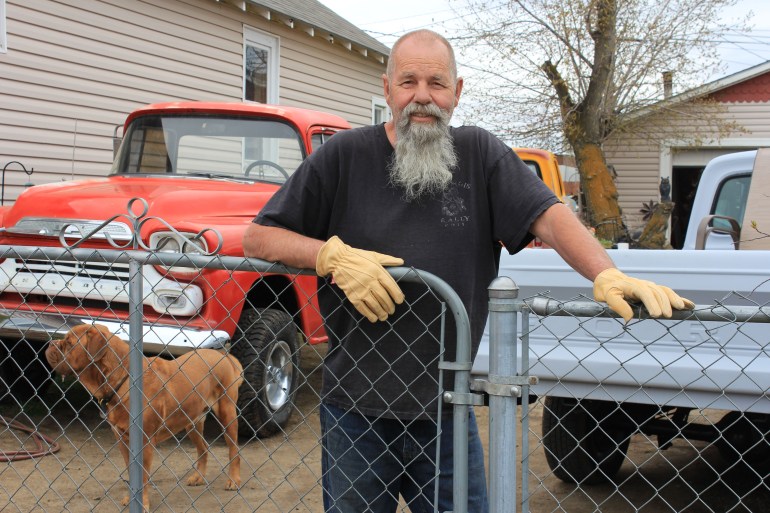 Al Shields stands in the center of the photograph behind a chain-link fence, which he rests his hands on. Behind him are two vintage trucks, one a soft red and the other pale blue.