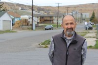 A photograph of Steve McGrath in the town of Butte, Montana. He looks directly at the camera and has his hands behind his back. Behind him are scattered buildings and beige cliffs.