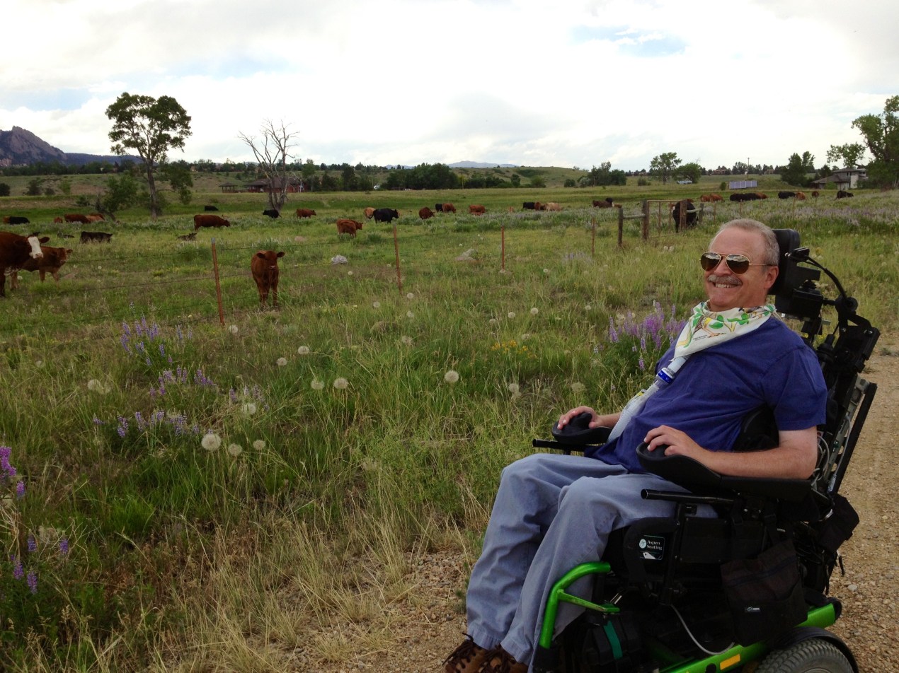 A photograph of Bruce Goguen sitting in his wheelchair outside, smiling at the camera. Beyond him is a sprawling pasture, where cows can be seen grazing.