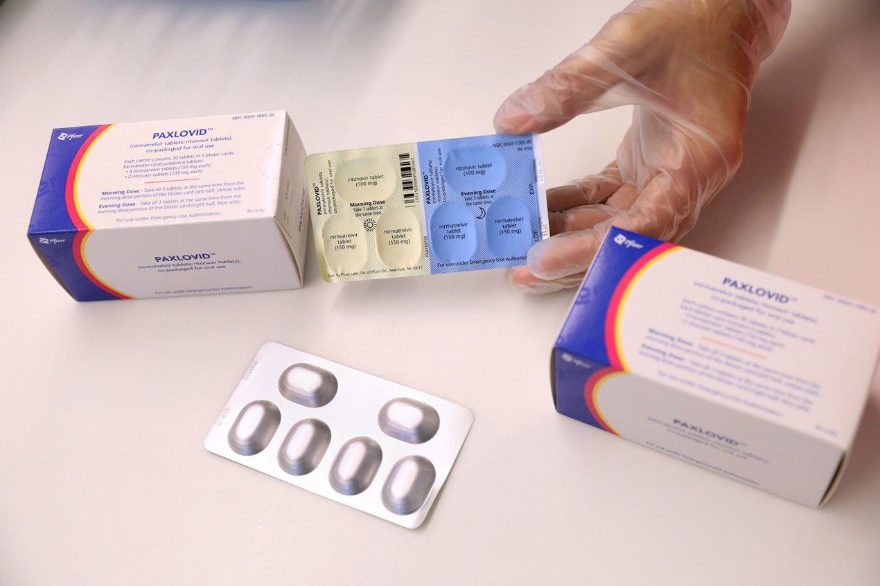 A photo shows a gloved hand holding a box of paxlovid. A blister pack of tablets is seen on the table.