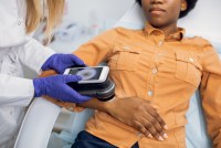 A photo shows a dermatologist holding a magnifier to a mole on a Black woman's wrist to check for melanoma.