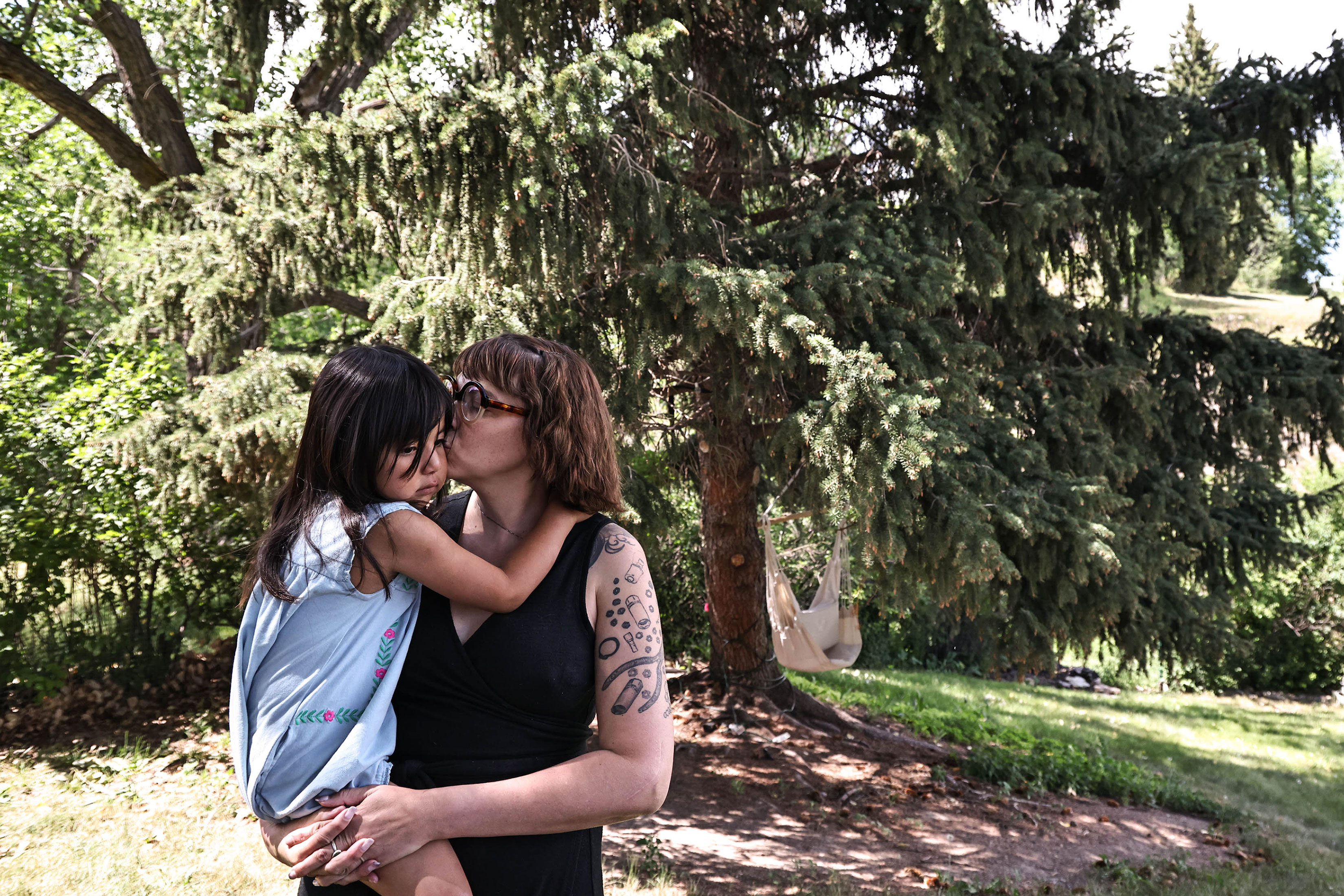 A woman holding a young child and kissing her on the cheek stands outside in front of a coniferous tree.