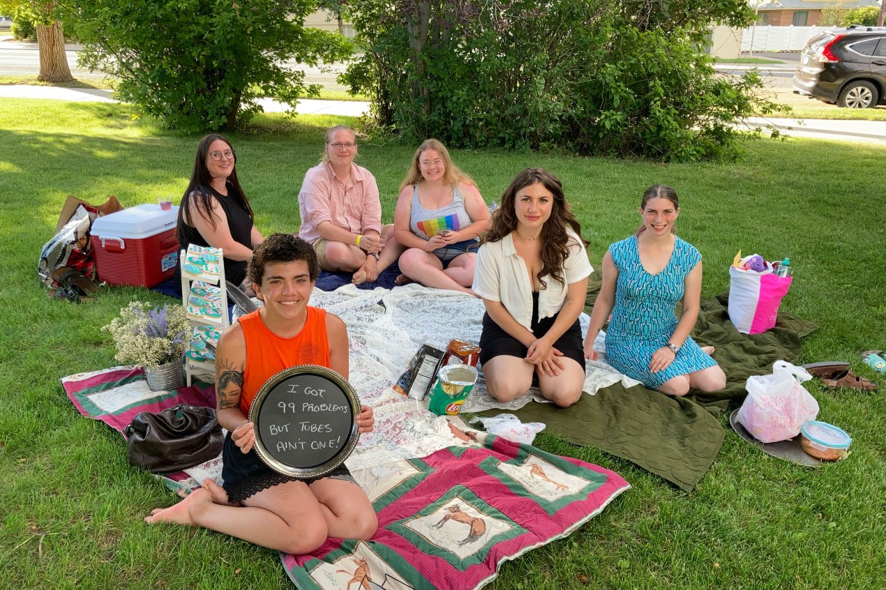 A photo shows Dani Marietti with her friends sitting on the grass set up for a picnic. She is holding a sign that reads, "I got 99 problems, but tubes ain't one."