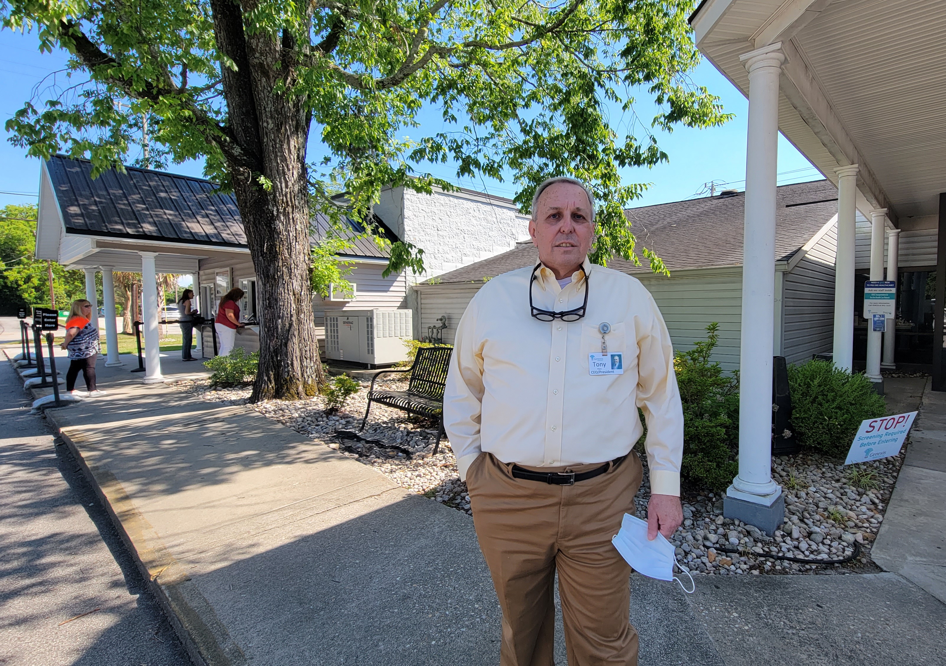 a man in a light shirt and slacks with glasses aroudn his neck stands nearby a white wooden building with a large tree in the background