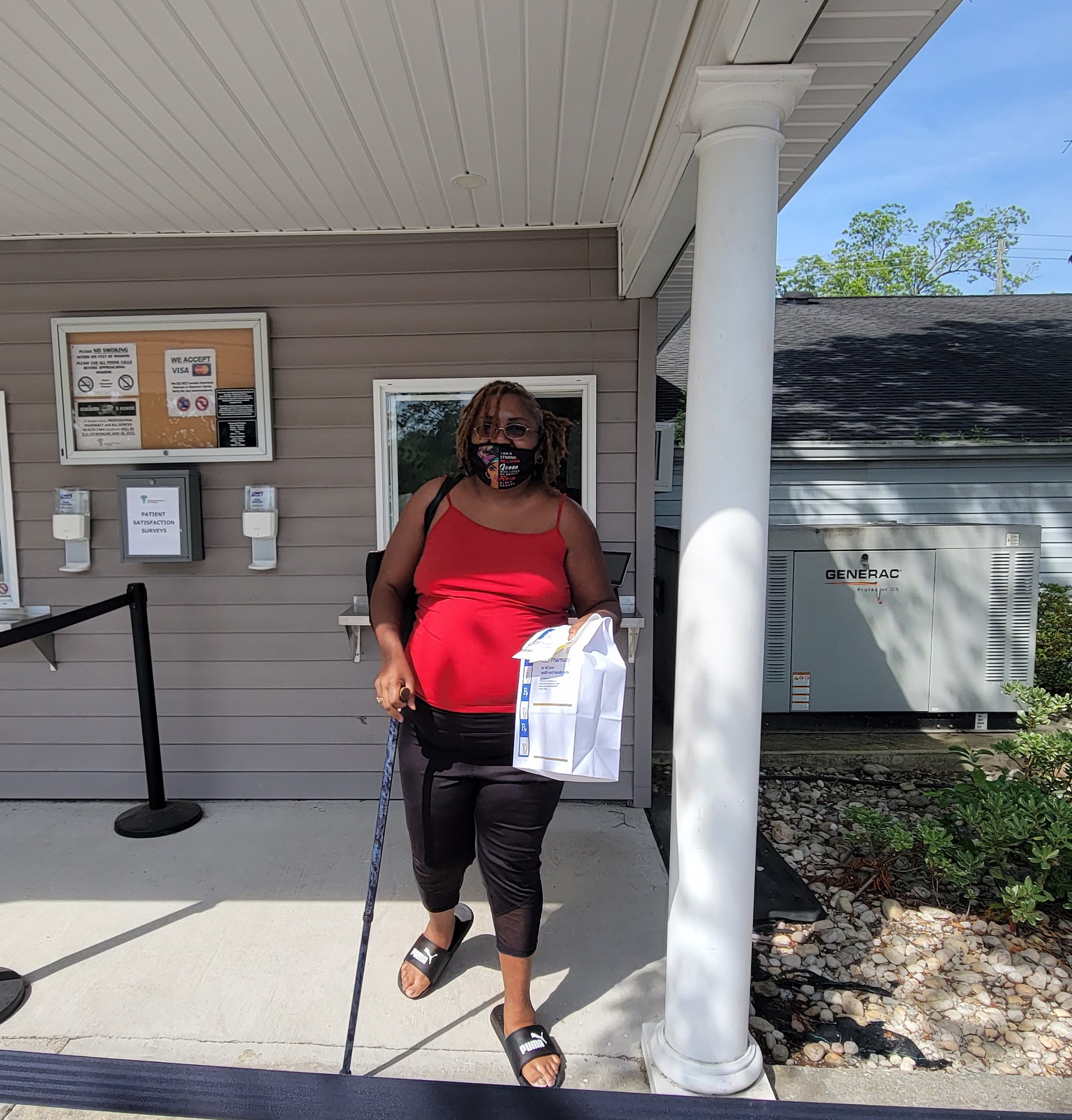 a woman in a red tank top with a walking stick stands outside a light colored wooden building with columns. she is holding a plastic bag in her left arm. there is a bulletin pinboard behind her on the building and a small customer service window