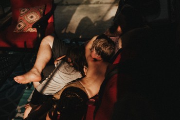 Two young boys that appear to be around 5 years old are draped over their father's lap, appearing exhausted. One of the boy's cheeks are flushed; he appears overheated. Sunlight comes in through a window, highlighting the family but casting the rest of the room in shadow.