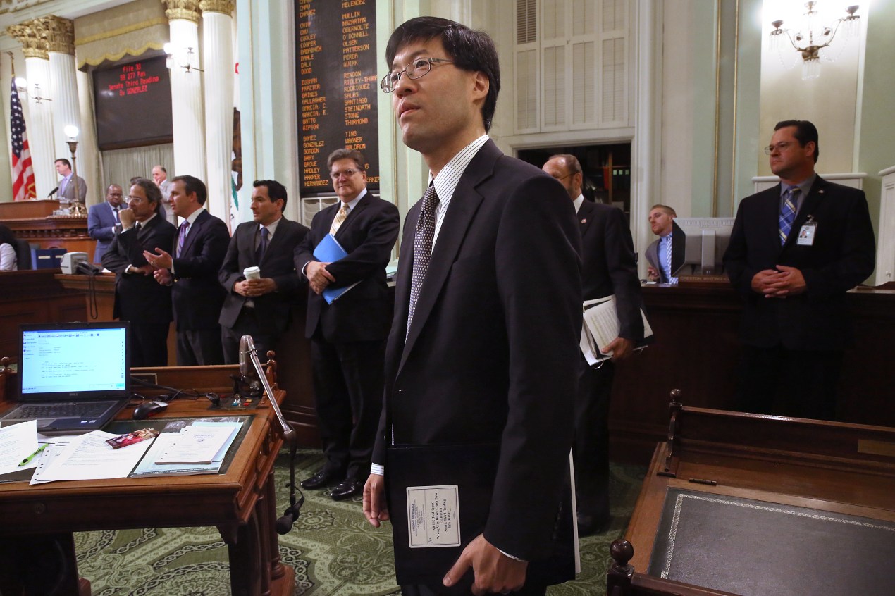 A man wearing a suit stands in a legislative chamber a looks at something out of frame.