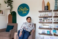 Tee Hundley, an adult female, sits in a chair in her nail salon. She has short, curly hair, wears large hoop earrings, a white blouse with a floral pattern, and jeans. The wall beside her holds nail products, and behind her is a sign that says, "Suite Tee".