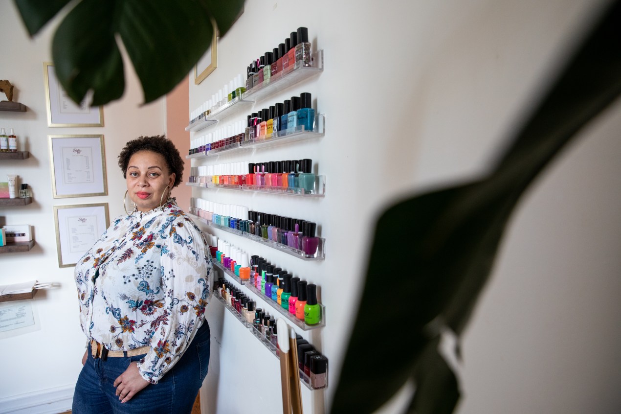 Tee Hundley leans up against a wall of colorful nail polish and looks directly towards the camera, with one hand in her pocket.