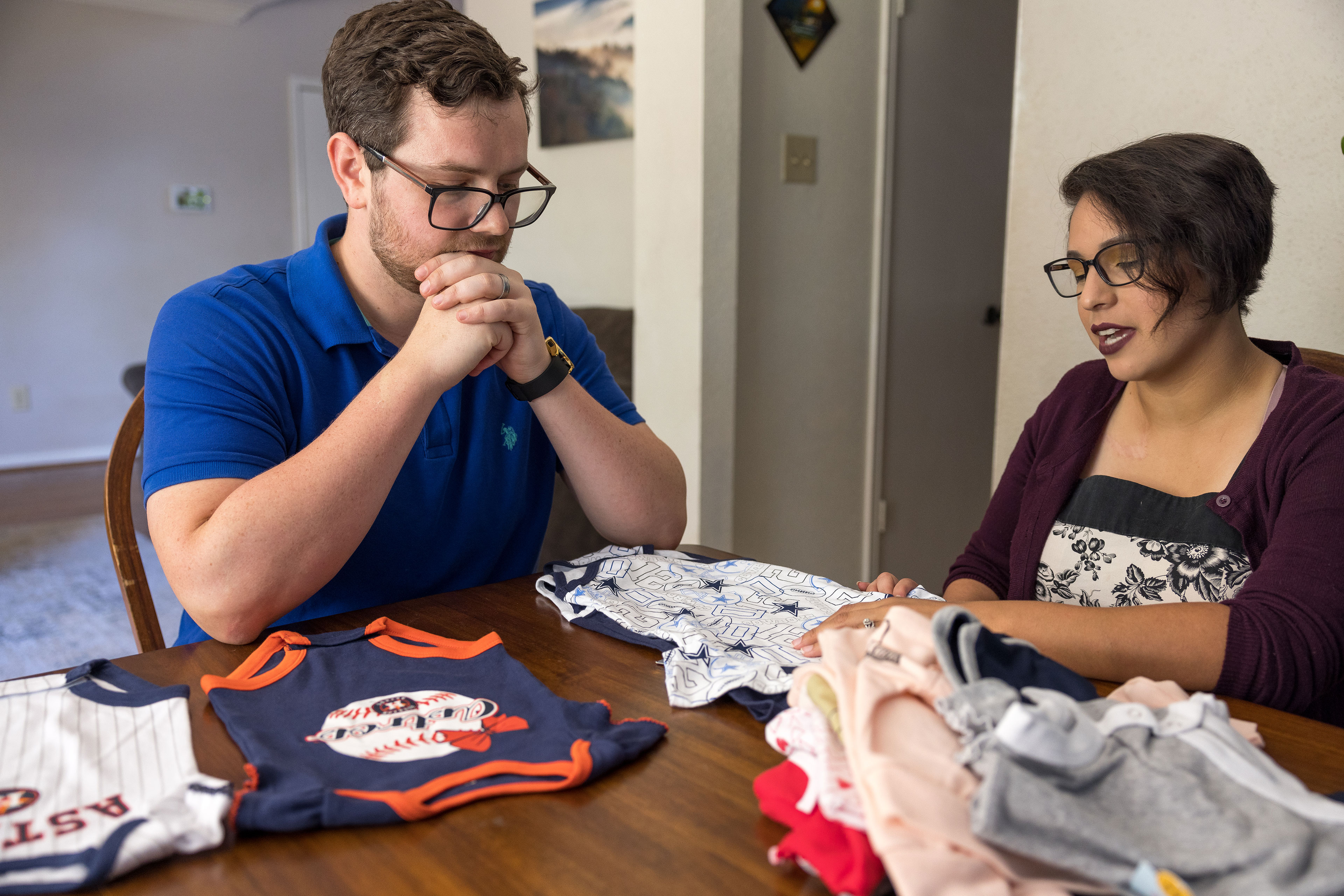 A man and a woman sit at a table where baby onesies are piled.