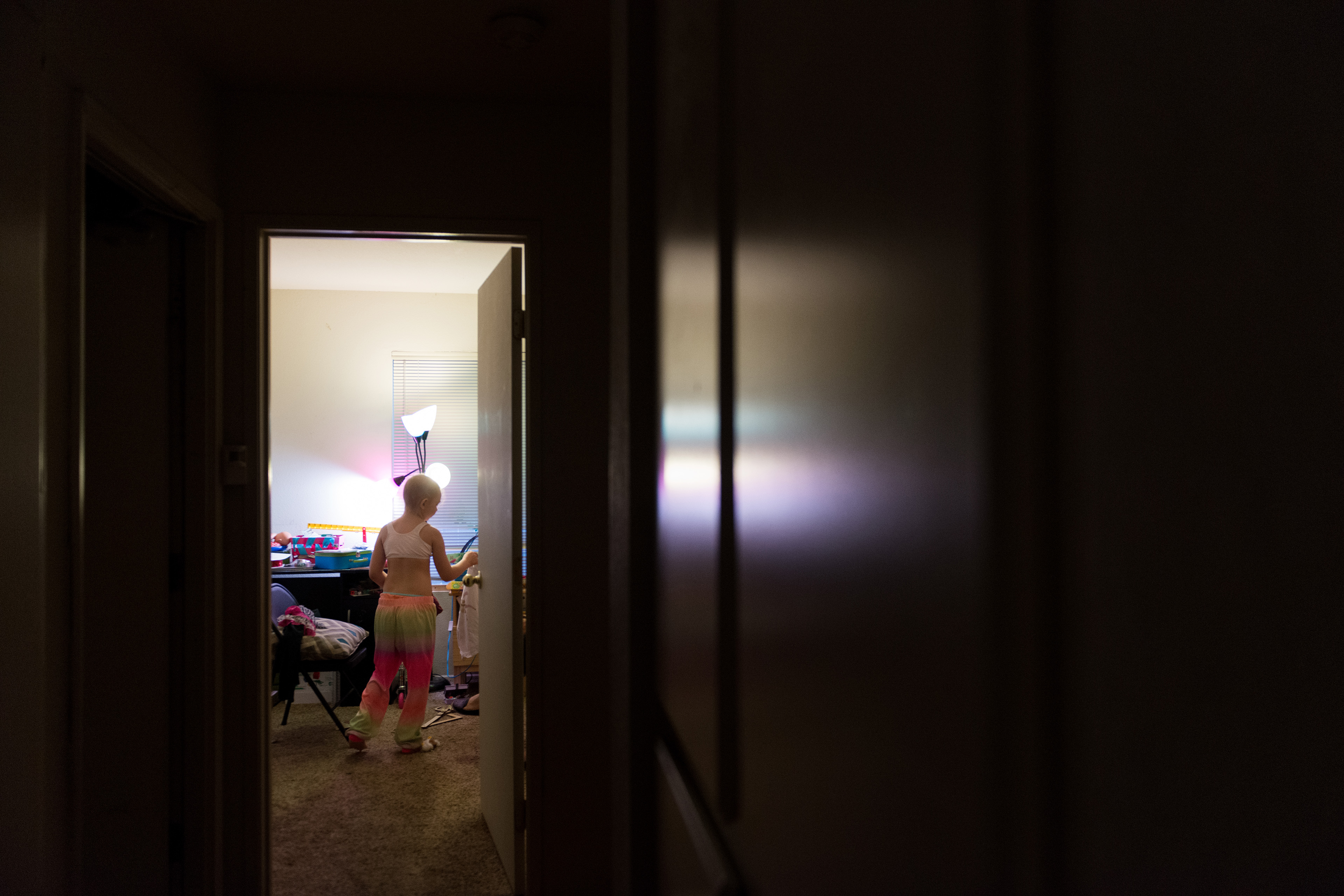 A doorway illuminates a long, dark hallway. In the room, you can see a young girl and some of her room, illuminated in a pinkish hue from her lamp.