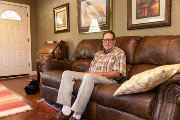 A man in a checkered shirt sits on a brown couch in a living room.