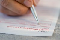A close-up photo shows someone filling out an insurance form with a pen.
