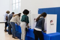 An unrecognizable mutli-ethnic group of voters stands to vote at the voting booths lined up against the wall of the gym.