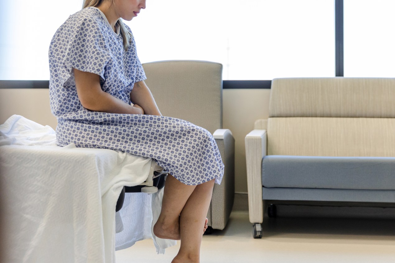 A photo shows a woman wearing a hospital gown and sitting at the edge of a hospital bed.