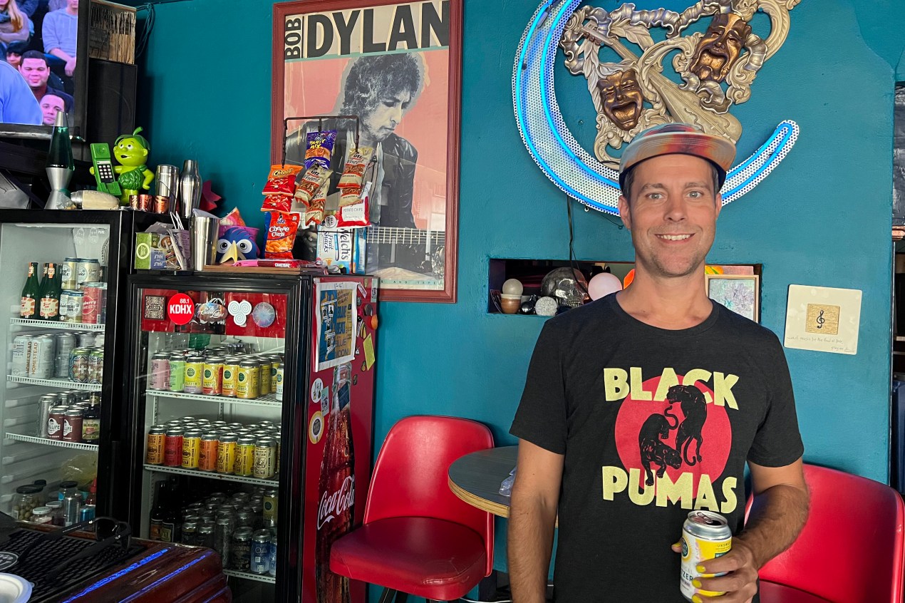 Joshua Grigaitis stands in his shop while holding a can of seltzer and smiling broadly. There are two mini-fridges visible behind him, each filled with seltzer and covered in stickers. A bright red chair and table are below a framed poster of Bob Dylan playing guitar. The walls are painted teal.