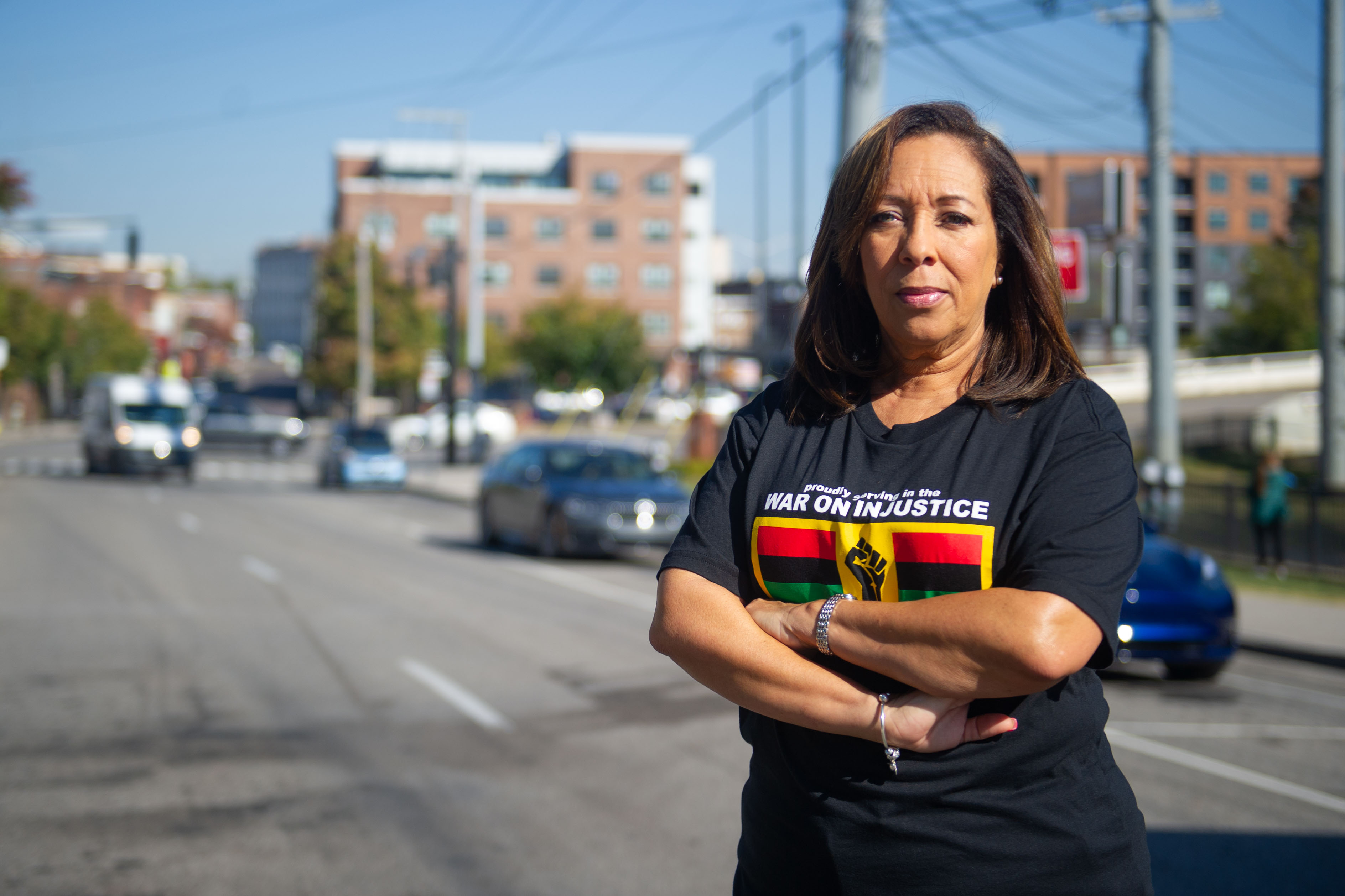 A Black woman wearing a T-shirt with the words "proudly striving in the WAR ON INJUSTICE" looks at the camera while standing beside a city street.