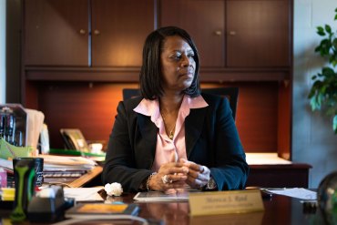 A Black woman in a light pink blouse and dark blazer sits behind a desk and looks off to her left.
