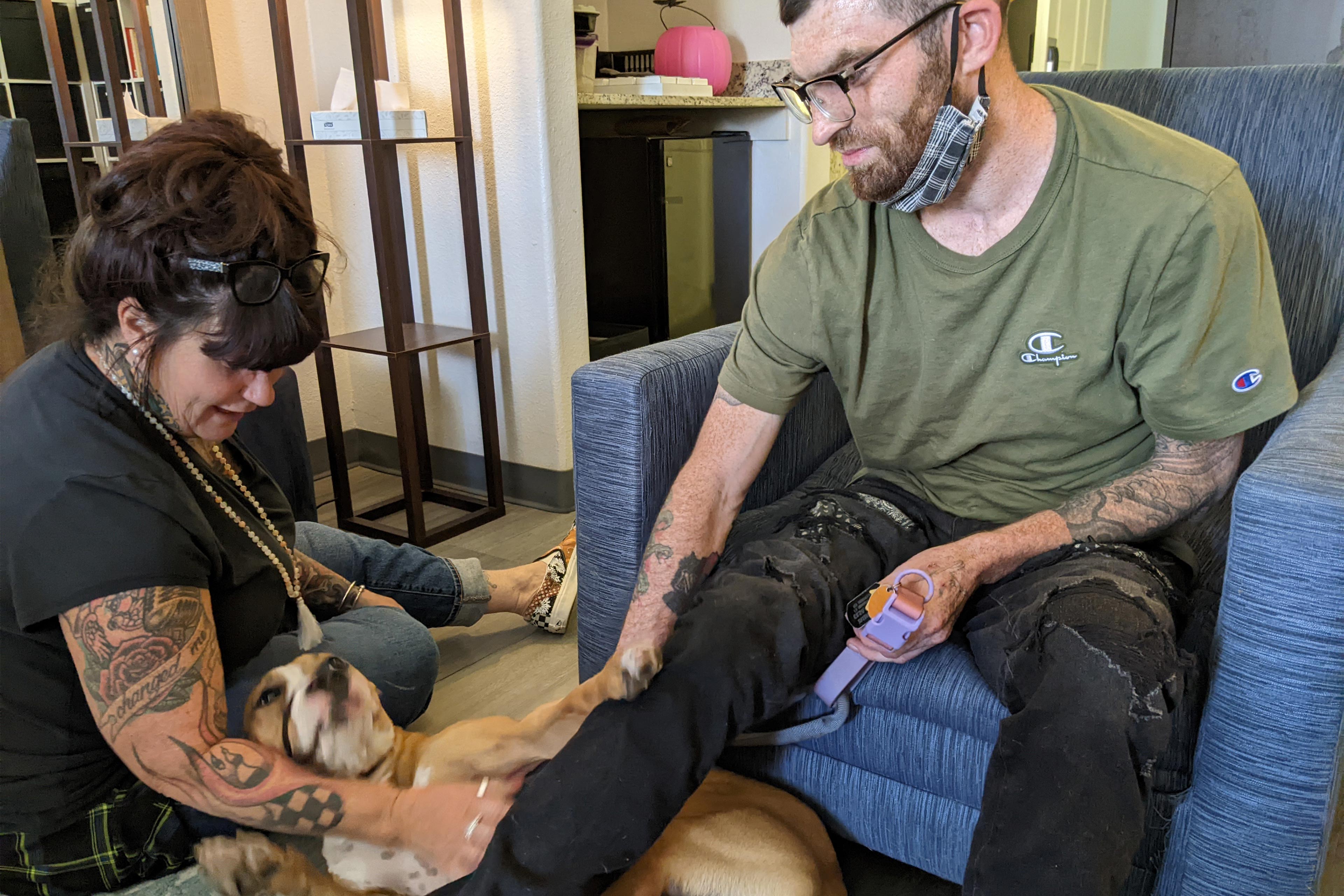 A photo shows Ian Dereus sitting in a chair as Donna Nortan pets his dog on the floor.