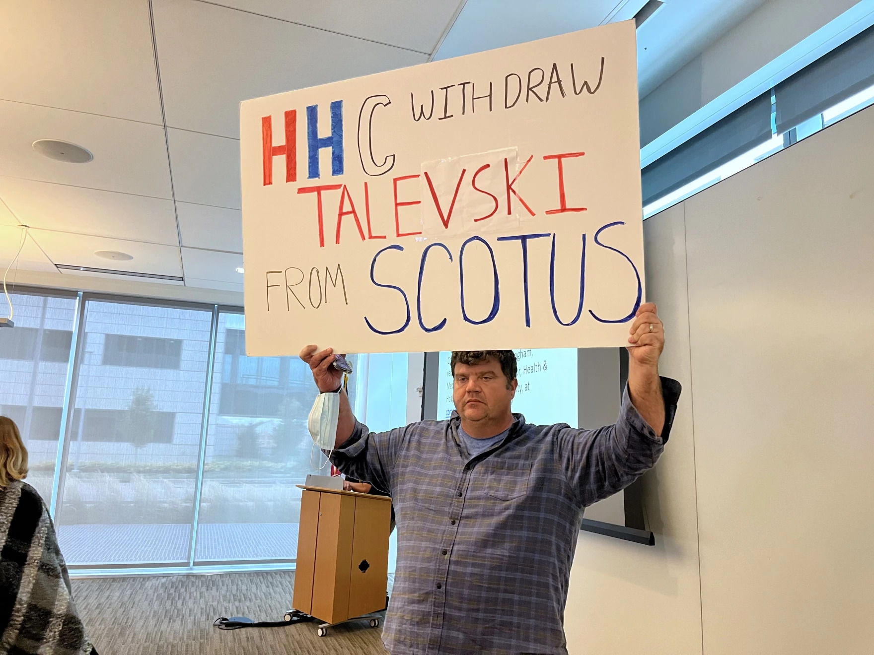A photo shows a man indoors holding up a sign that reads, "HHC withdraw Talevski from SCOTUS."
