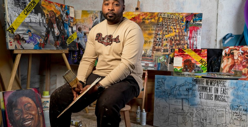 A photo shows Mykael Ash sitting in his studio, surrounded by his paintings behind him. He's holding a paintbrush and palette in his hands.