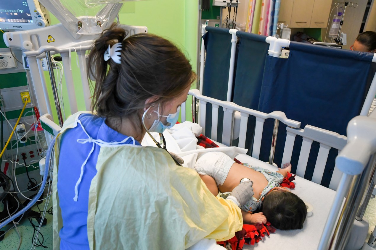 A hospital staff member is caring for an infant in a pediatric care unit, which resembles a crib. The infant is laying on its stomach with its head turned away from the camera. 