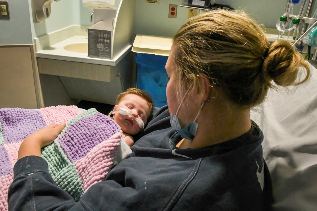 Caitlyn Houston holds her infant daughter, Parker, as they wait in the emergency department. The photo is taken from over the shoulder of the mother, which reveals the infant's face. The infant has an oxygen tube on her face, below the nose. She is sleeping.