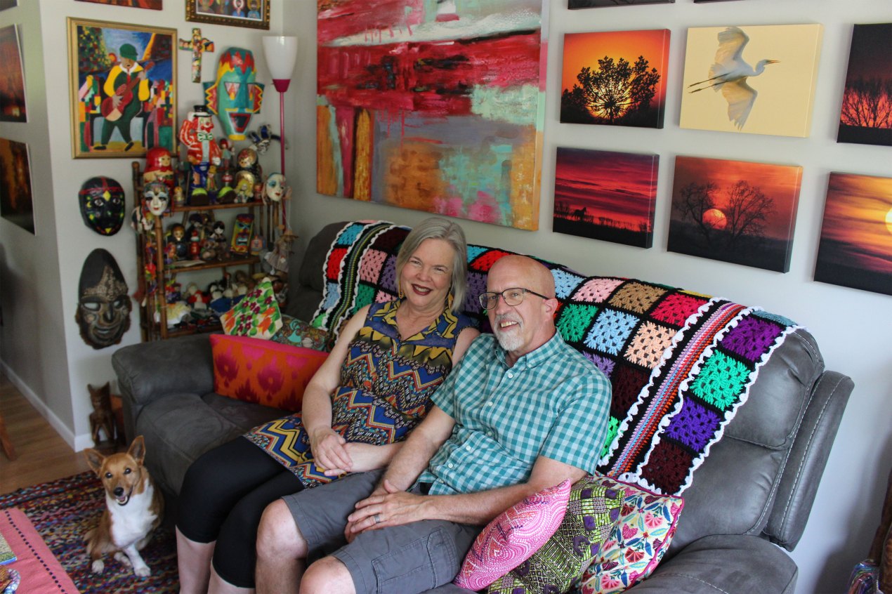 A woman and man sit on a dark gray couch covered in several colorful pillows and a crocheted blanket. Colorful artwork hangs on the walls behind and beside the couch and a small tan and white dog sits on the floor besides the couple.