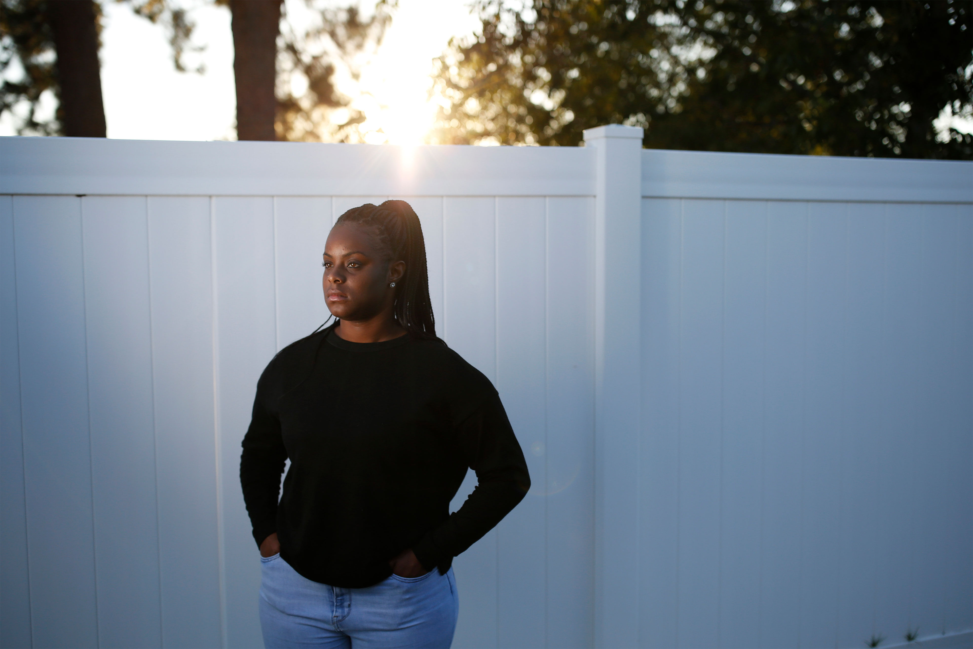 A woman stands outside in front of a tall white fence in late afternoon sun.