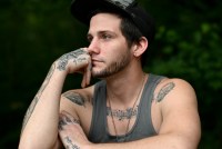 Adam Caprioli sits with one arm crossed over his chest while he rests his cheek against his opposite hand. He is gazing off to the side with a distant, but thoughtful expression, like trying to recall a memory.